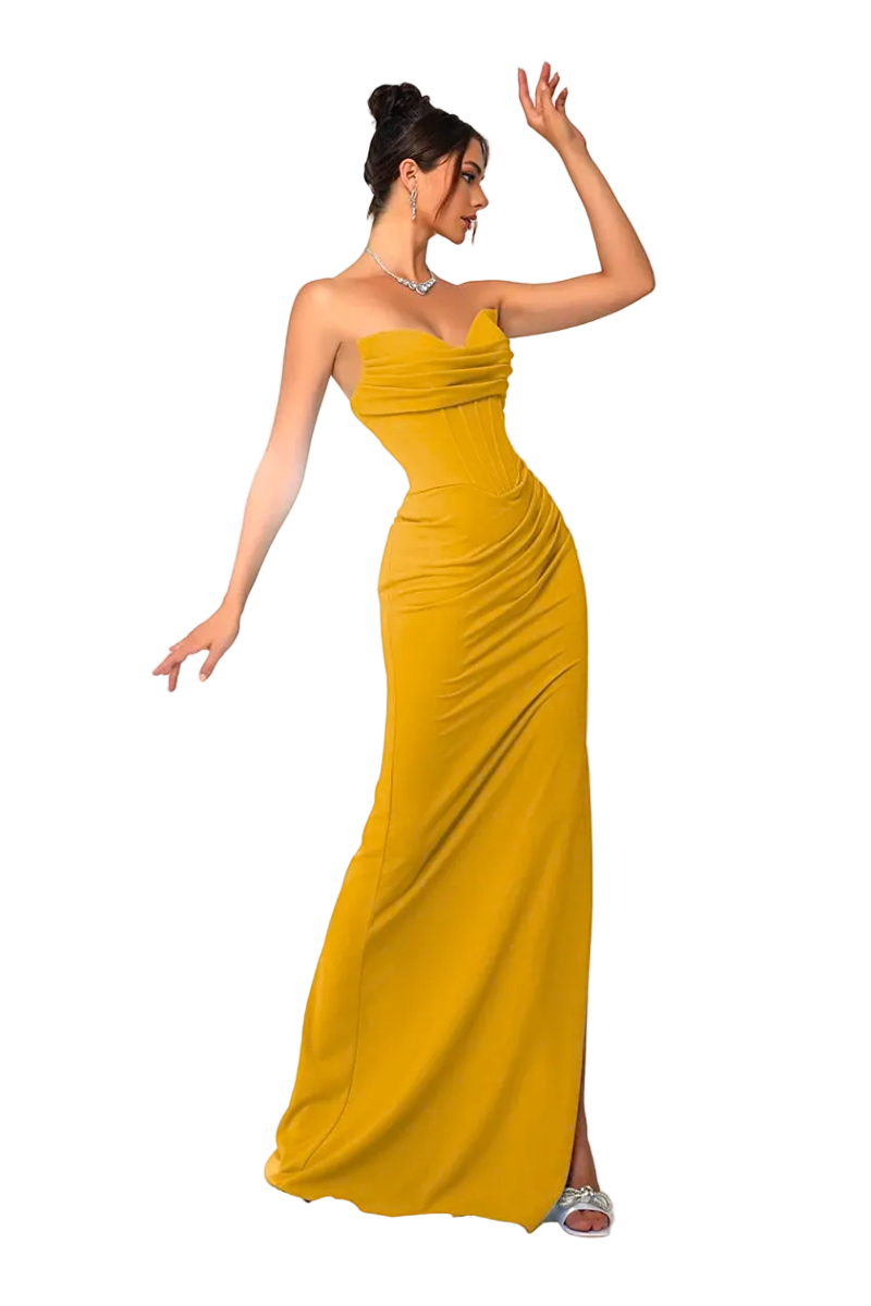 A woman models a strapless mustard yellow floor-length gown with ruching and a side slit, posed in front of white draped fabric with a white pedestal and bird prop.