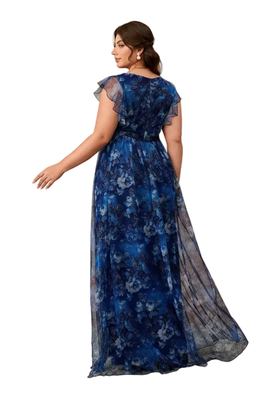 A woman modeling a floor-length blue floral gown with sheer cap sleeves and a sheer side panel, photographed in a studio with warm beige arched walls.