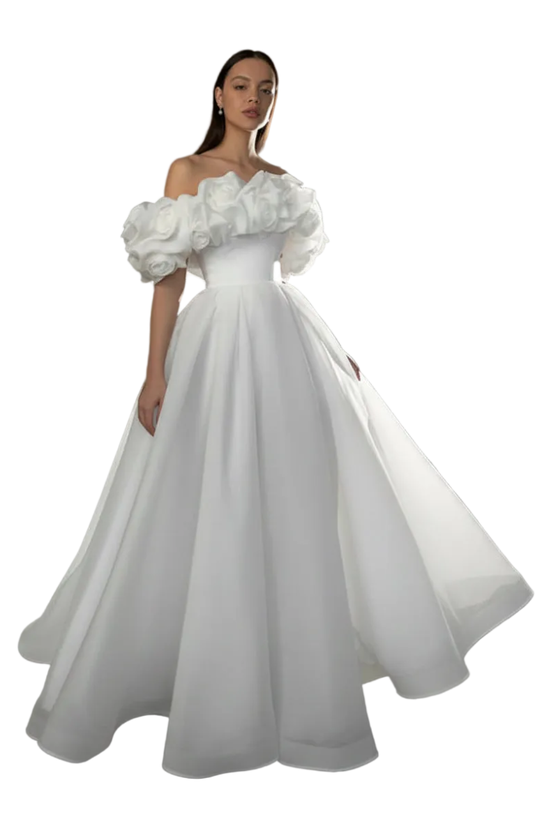 A woman modeling a white off-the-shoulder ball gown with dramatic puffed sleeves and a full skirt, photographed in a studio setting with a gray backdrop and a bright light fixture visible in the background.