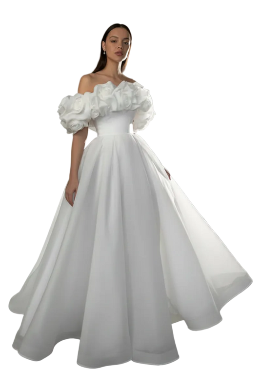 A woman modeling a white off-the-shoulder ball gown with dramatic puffed sleeves and a full skirt, photographed in a studio setting with a gray backdrop and a bright light fixture visible in the background.