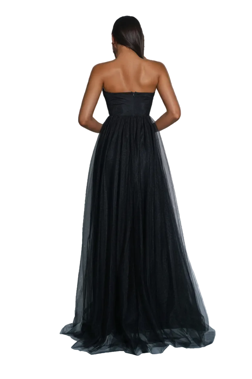 A woman stands with her back to the camera wearing a strapless black formal gown with a fitted bodice and a voluminous tulle skirt. The photo is taken in a studio with gray walls and floor, with a silver disco ball on the left.