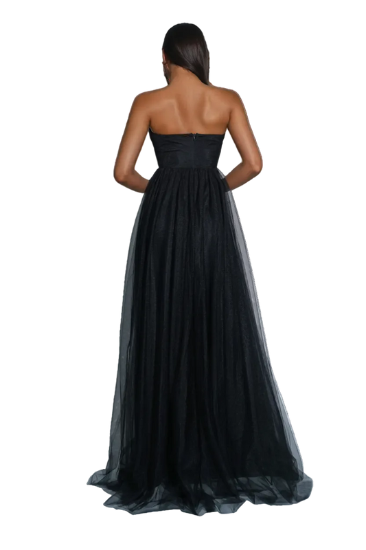 A woman stands with her back to the camera wearing a strapless black formal gown with a fitted bodice and a voluminous tulle skirt. The photo is taken in a studio with gray walls and floor, with a silver disco ball on the left.