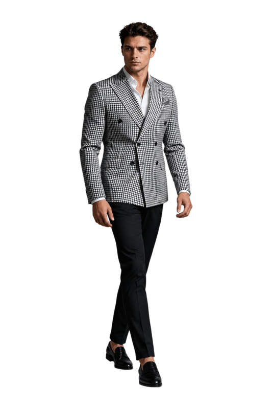 A man modeling a double-breasted blazer with a black and white houndstooth pattern, paired with a white dress shirt and black trousers, set against a textured gray studio background with a 'UA' logo in the top left corner.