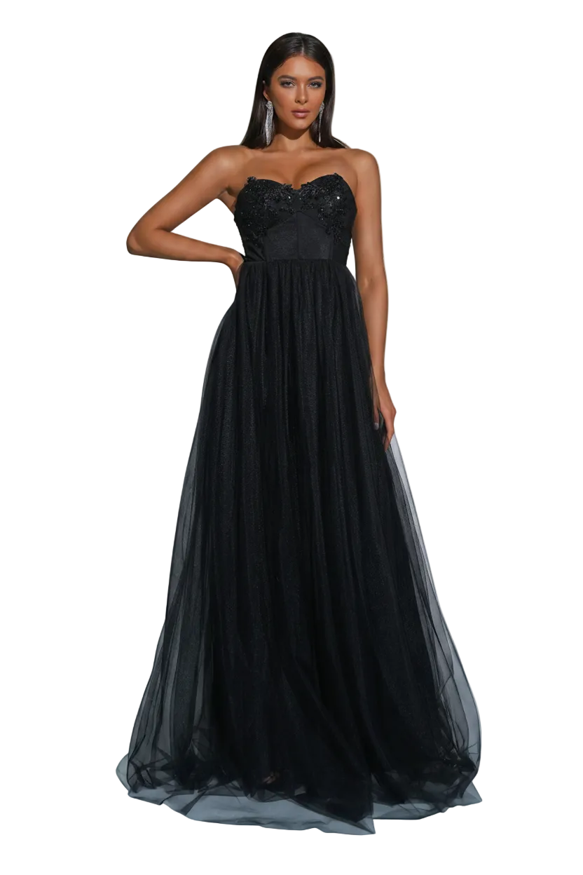 A woman models a strapless black formal gown with a sweetheart neckline, embellished bodice, and a voluminous tulle skirt. The image is set in a studio with gray walls, spotlighting, and decorative silver disco balls on the floor.